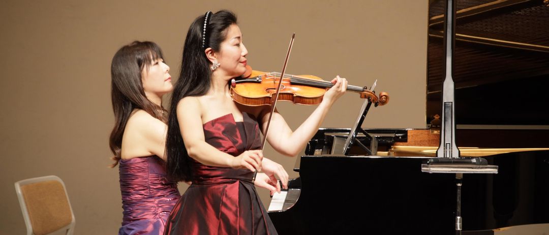 Zwei Frauen, gekleidet in elegante Kleider, spielen auf der Bühne. Eine spielt Geige, während die andere am Klavier sitzt.