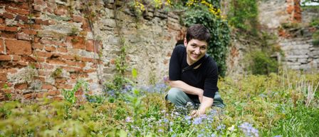 Eine Person kniet in einem Feld mit blauen Blumen und lächelt. Hinter ihr ist eine Backsteinmauer, die mit Grün und gelben Blumen bedeckt ist.