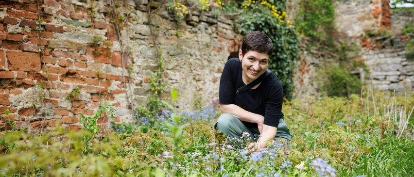 Eine Person kniet in einem Feld mit blauen Blumen und lächelt. Hinter ihr ist eine Backsteinmauer, die mit Grün und gelben Blumen bedeckt ist.