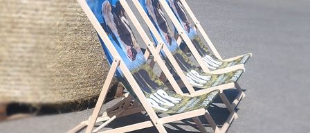 Three wooden folding chairs with printed images of people are arranged in a row on a concrete surface. The chairs are blue and green.