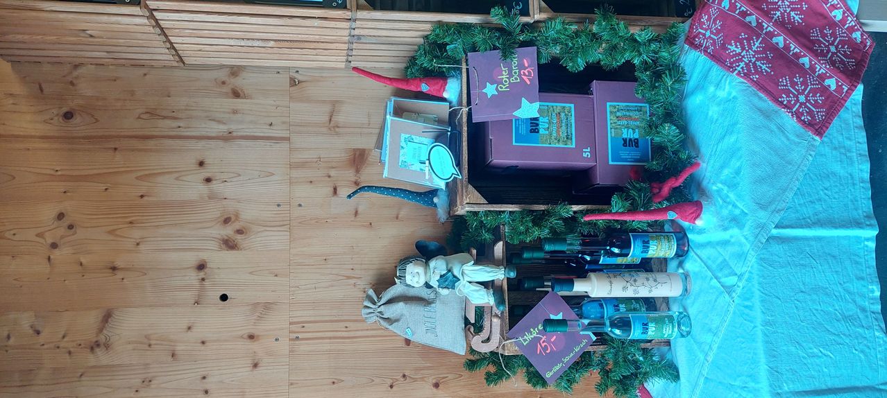 Ein Holzregal zeigt verschiedene Gegenstände, darunter Weihnachtsdekorationen, Bücher, Flaschen und Figuren. Die Artikel sind sorgfältig arrangiert und mit Etiketten versehen, die Text und Preise enthalten.