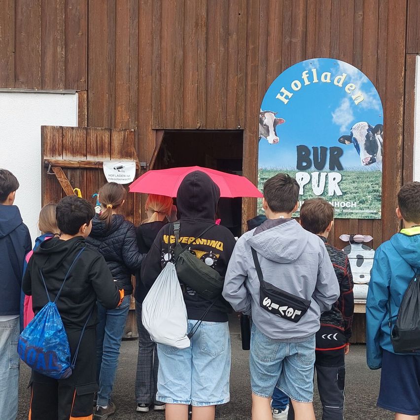A group of children stands outside a building with a blue sign that reads 'Hoflladen BUR PUR' with cows on it. One child holds a red umbrella.