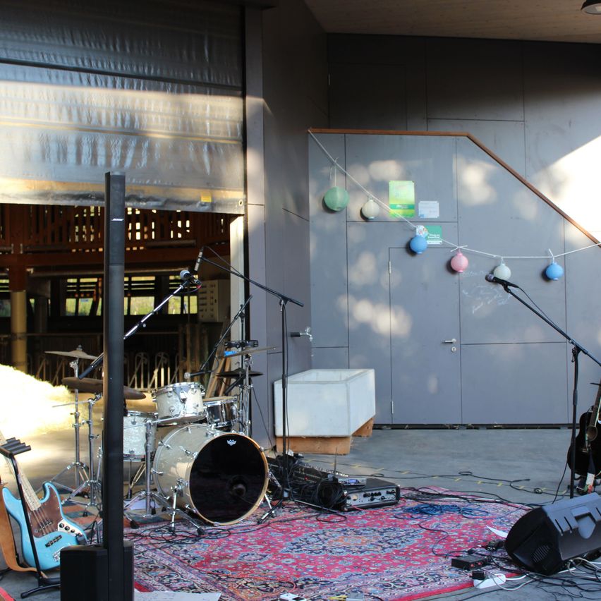 A drum set, microphones, and various musical instruments are set up on a stage with a rug, speakers, and a staircase in the background.