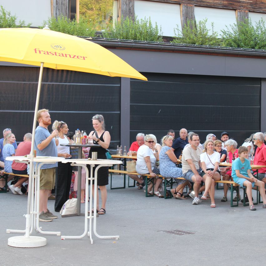 A group of people are seated at a beer garden, with two servers standing behind tables under a yellow umbrella. The garden is surrounded by a building with a green roof.