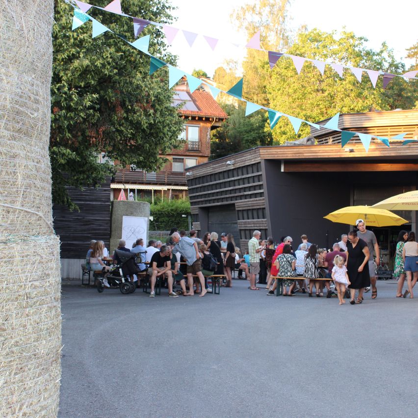 A group of people are gathered in an outdoor area, some sitting on benches while others are walking around. The area is decorated with bunting and there are buildings in the background.