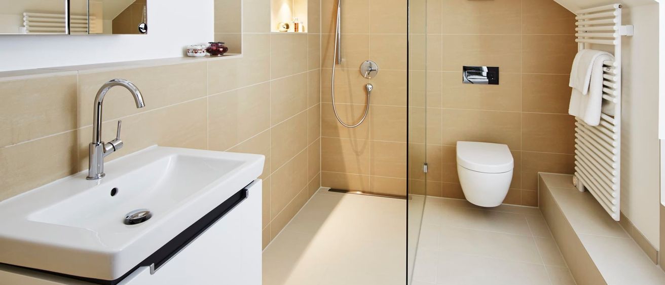 Ein modernes Badezimmer mit beigen Fliesen, einer Glasdusche, einem weißen WC, einem silbernen Wasserhahn und einem Spiegel an der Wand.