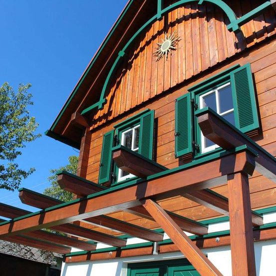 Ein Holzhaus mit einem Sonnendesign über den Fenstern, grünen Fensterläden und einer Holzpergola.