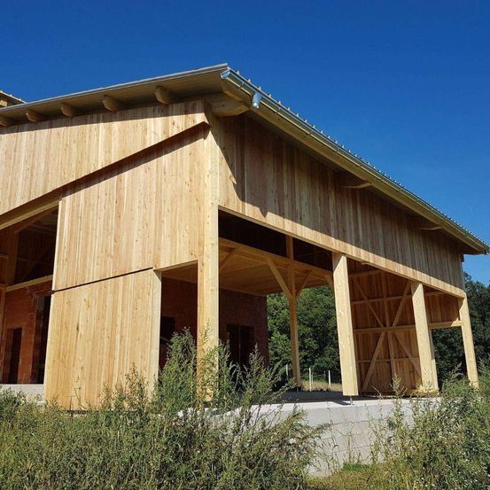 Ein Holzschuppen im Bau mit einem geneigten Dach und großen Holzbalken, die die Struktur stützen. Er ist von Grün umgeben.