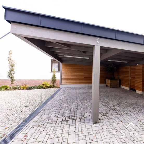 Ein Carport mit grauem Dach, gestützt von Holzsäulen, bedeckt eine gepflasterte Einfahrt. Ein Topf mit Pflanzen steht rechts.