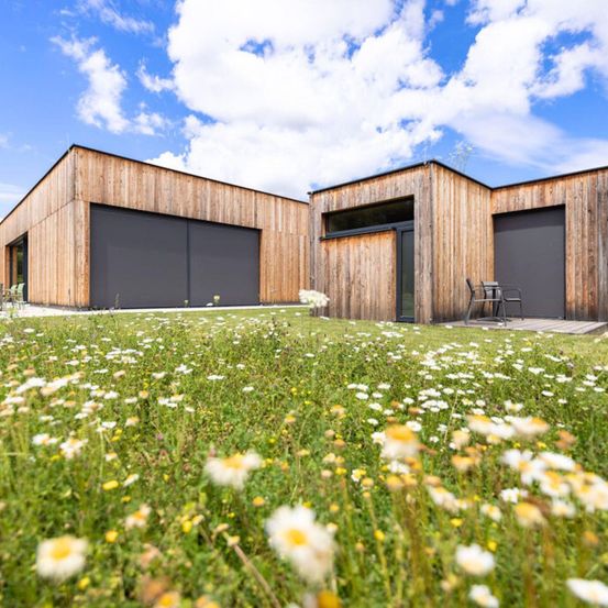 Ein modernes Holzhaus mit zwei Garagen und einer Tür, in einer üppigen, blumenreichen Wiese unter einem teilweise bewölkten Himmel.