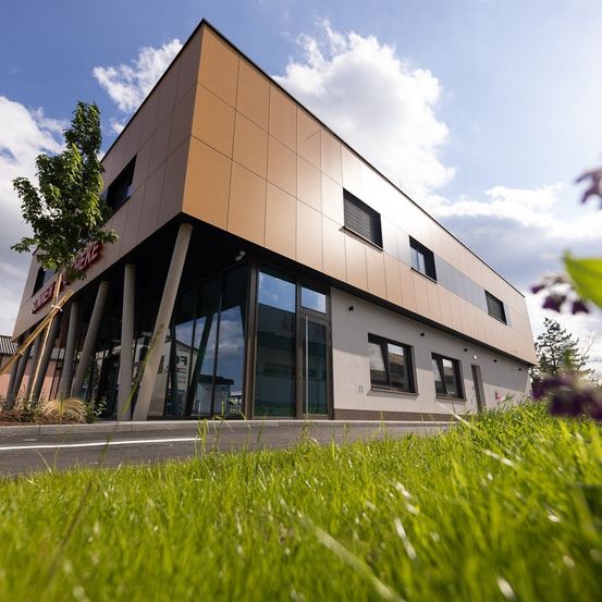Ein modernes Gebäude mit brauner Außenfassade und Glasfenstern, in einem grünrasigen Bereich gelegen. Der Himmel ist blau mit einigen Wolken.