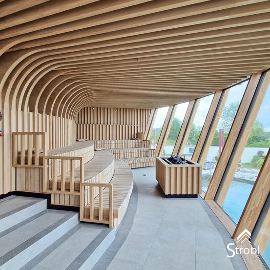 Das Bild zeigt einen modernen Spa-Bereich mit Holzbänken und einer gebogenen Decke. Große Fenster bieten Blicke auf einen Pool im Freien. Ein hölzerner Pflanzkasten steht in der Nähe der Glaswände.