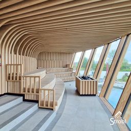Das Bild zeigt einen modernen Spa-Bereich mit Holzbänken und einer gebogenen Decke. Große Fenster bieten Blicke auf einen Pool im Freien. Ein hölzerner Pflanzkasten steht in der Nähe der Glaswände.