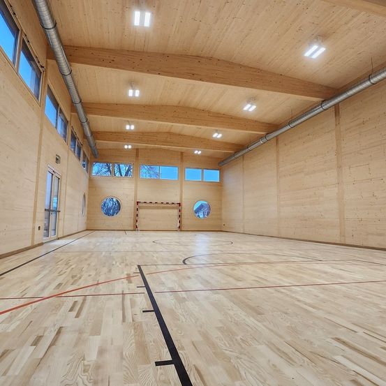Eine leere Indoor-Sporthalle mit Holzwänden und -böden, ausgestattet mit einem Fußballtor und großen Fenstern mit runden Öffnungen. Die Decke ist aus Holz mit Metallrohren und Einbauleuchten.