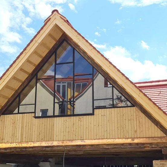 Ein dreieckiges Gebäude mit Holzfassade und rotem Dach. Es hat große Glasfenster mit schwarzen Rahmen. Der Himmel ist blau mit verstreuten Wolken.