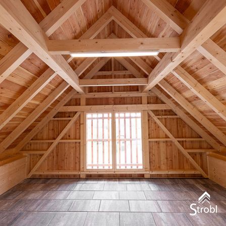 Ein leerer Dachboden mit Holzbalken, Bodenbelag und einem Glasfenster. Ein Logo ist auf dem Boden sichtbar.