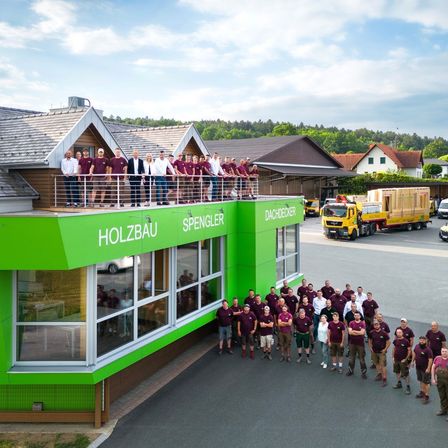 Eine Gruppe von Menschen in maroon Uniformen posiert für ein Foto vor einem grünen Gebäude mit 'Holzbau' und 'Spengler Dachdecker' Schildern. Im Hintergrund befinden sich Häuser, Bäume und ein gelber Lastwagen.