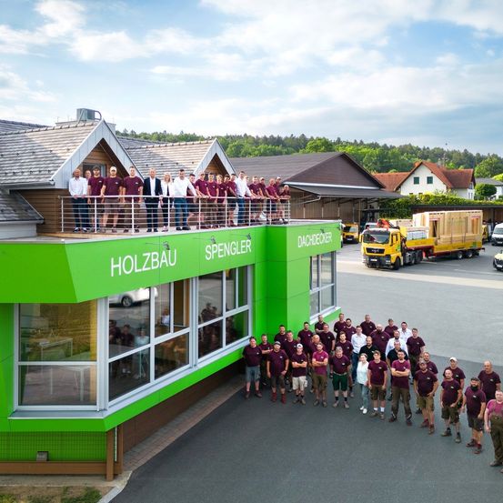Eine Gruppe von Menschen in maroon Uniformen posiert für ein Foto vor einem grünen Gebäude mit 'Holzbau' und 'Spengler Dachdecker' Schildern. Im Hintergrund befinden sich Häuser, Bäume und ein gelber Lastwagen.