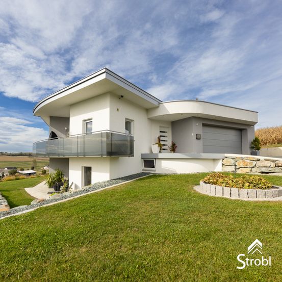 Ein modernes weißes Haus mit Glasbalkon und Garage, in einer Rasenlandschaft mit Steinmauer und Pflanzen.