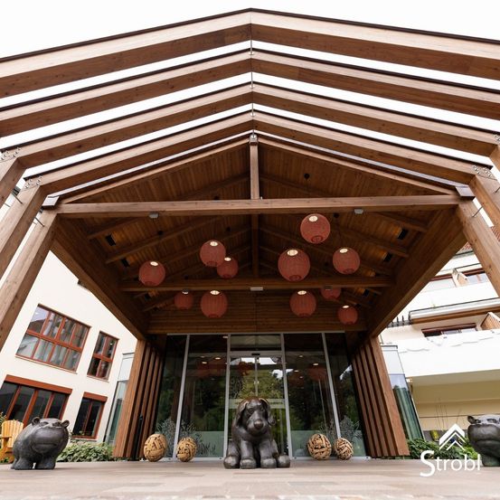 Eine Holzstruktur mit einem geometrischen Dachdesign. Rote Laternen hängen von der Decke. Eine Statue eines Hundes und dekorative Bälle stehen vor dem Eingang auf dem Boden.