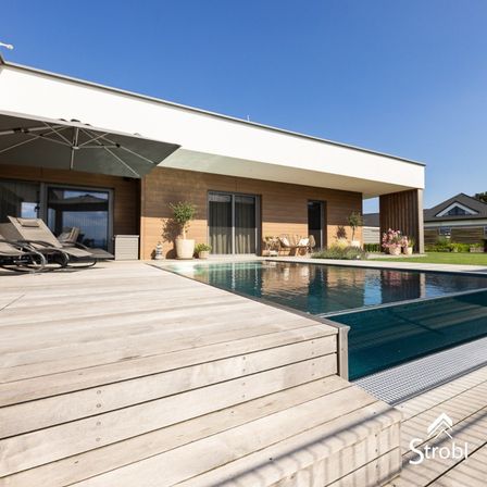 Ein modernes Haus mit Holzterrasse, einem Regenschirm, Liegestühlen und einem Pool mit Blick auf das Meer.