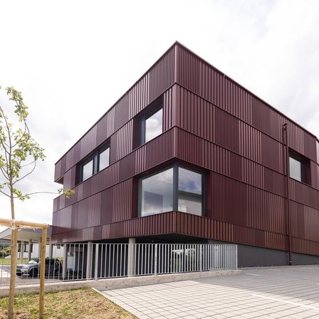 Ein modernes Gebäude mit roten Metallplatten und großen Fenstern. Ein Auto ist vor einem Metallzaun geparkt.