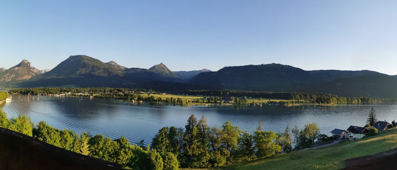 Ein friedlicher Landschaftsblick auf einen großen See, umgeben von grünen Bäumen, einem grasbewachsenen Hügel und Bergen unter einem klaren Himmel.