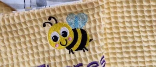 Eine gelbe Decke mit Bienenstickerei darauf und dem Wort Theres. Darunter eine kleinere Decke mit der gleichen Stickerei und dem Wort.