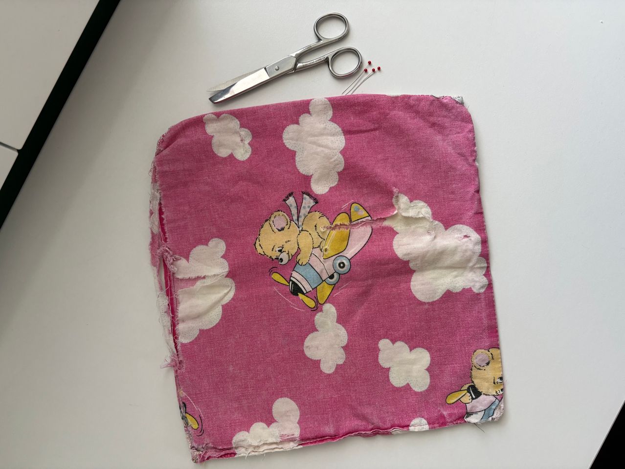 Ein rosa Stoff mit einem fliegenden Bären in einer Rakete liegt auf einer weißen Oberfläche. Daneben sind Schere und Nadeln.