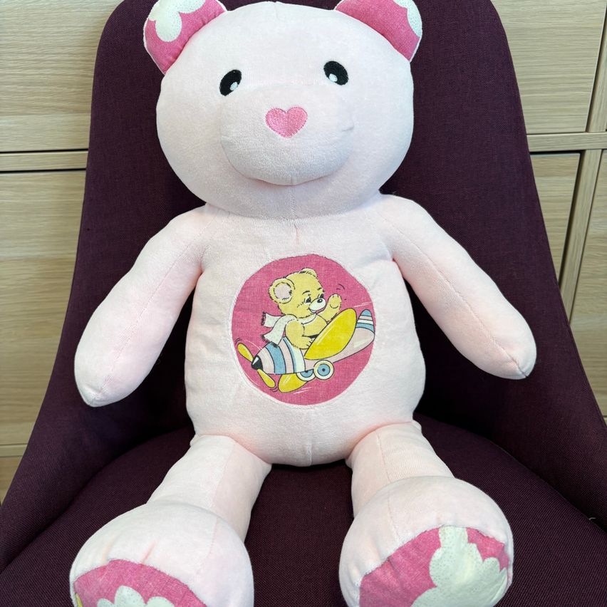 Ein rosa Plüsch-Teddybär sitzt auf einem lila Stuhl. Er hat ein rosa Herz auf der Nase und einen runden Aufkleber auf der Brust. Der Aufkleber zeigt einen Cartoon-Bären, der einen Skateboard fährt.