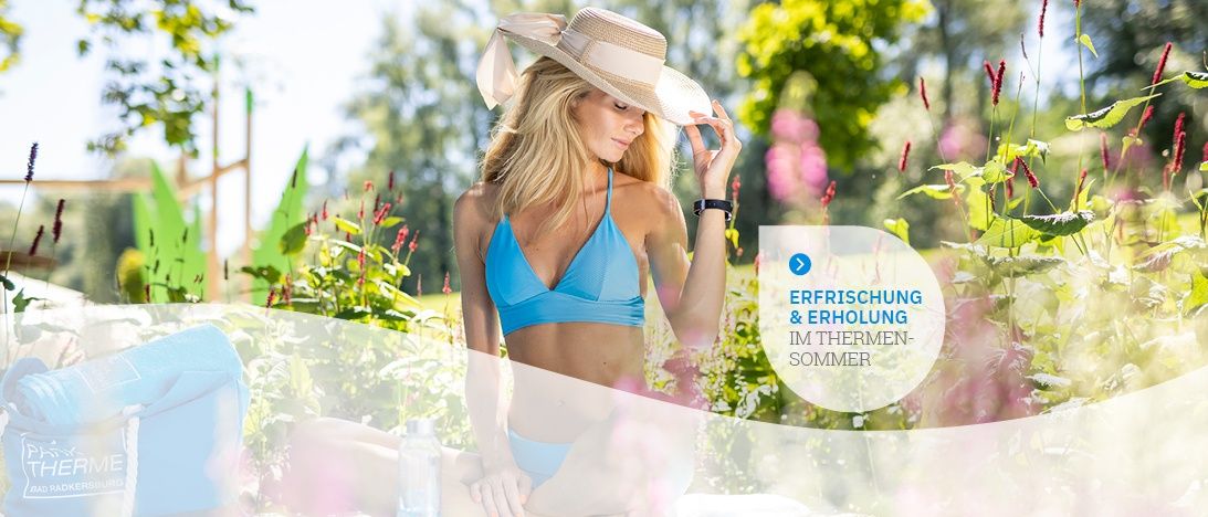 Eine Frau in einem blauen Bikini und Hut sitzt auf einer Bank in einem Garten. Sie hält ihren Hut und schaut etwas an. Hinter ihr ist ein unscharfes Bild einer Person, die auf dem Boden sitzt.