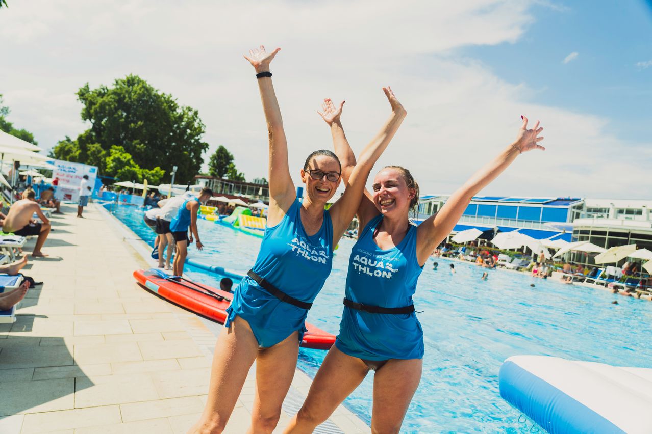 Zwei Frauen in blauen Badeanzügen und Aquathlon-Shirts feiern in einem Freibad, wobei andere Schwimmer und Aufblasbare im Hintergrund zu sehen sind.