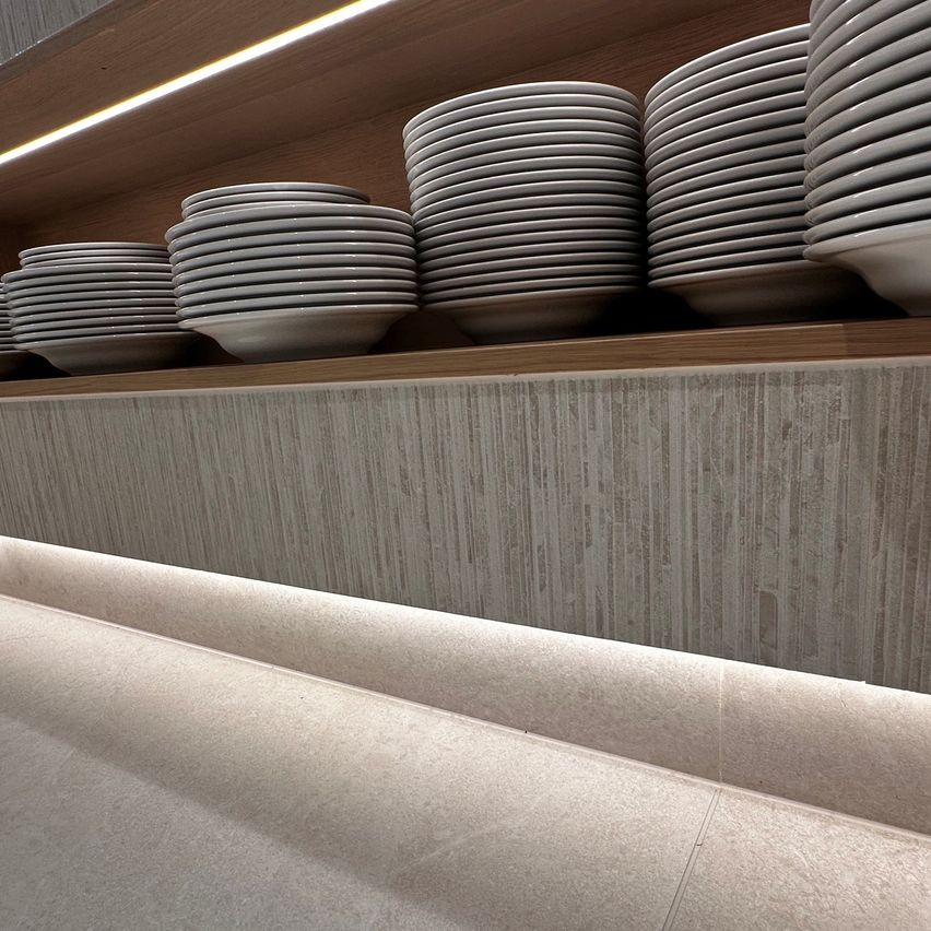 A wooden shelf with multiple stacks of white plates neatly arranged, illuminated by a soft light. The background is a textured gray wall.