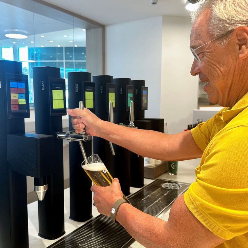 An older man with glasses and a yellow shirt is smiling while pouring a glass of beer from a tap in a bar.