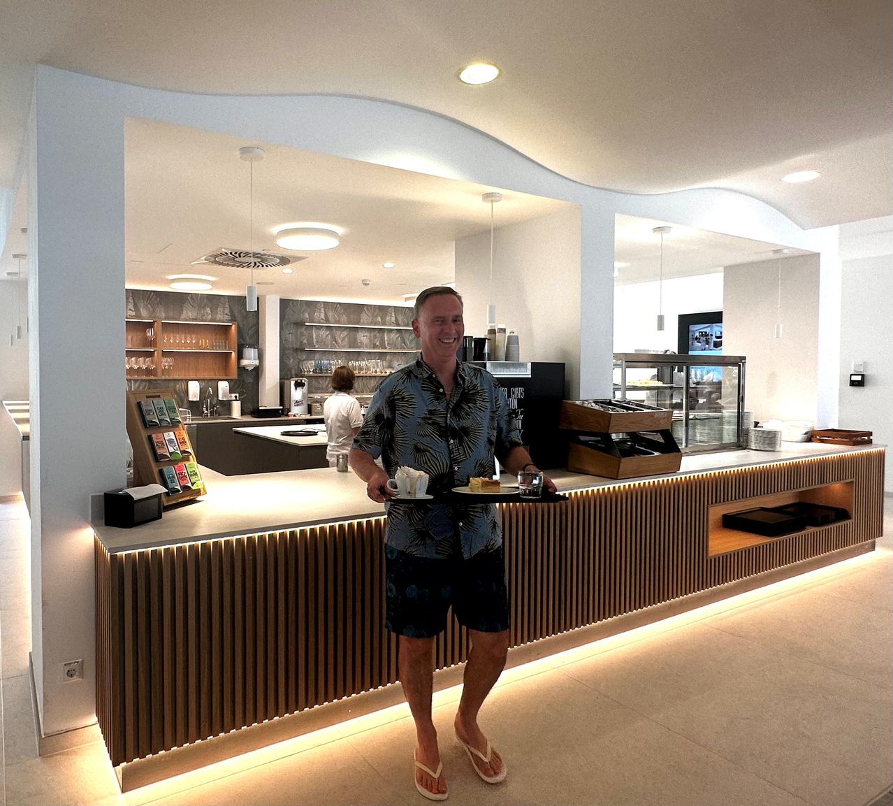A man in a floral shirt and shorts stands behind a counter, smiling and holding a tray with a cup and saucer, and a plate of food. Behind him, a woman stands at a counter with shelves and a coffee machine.