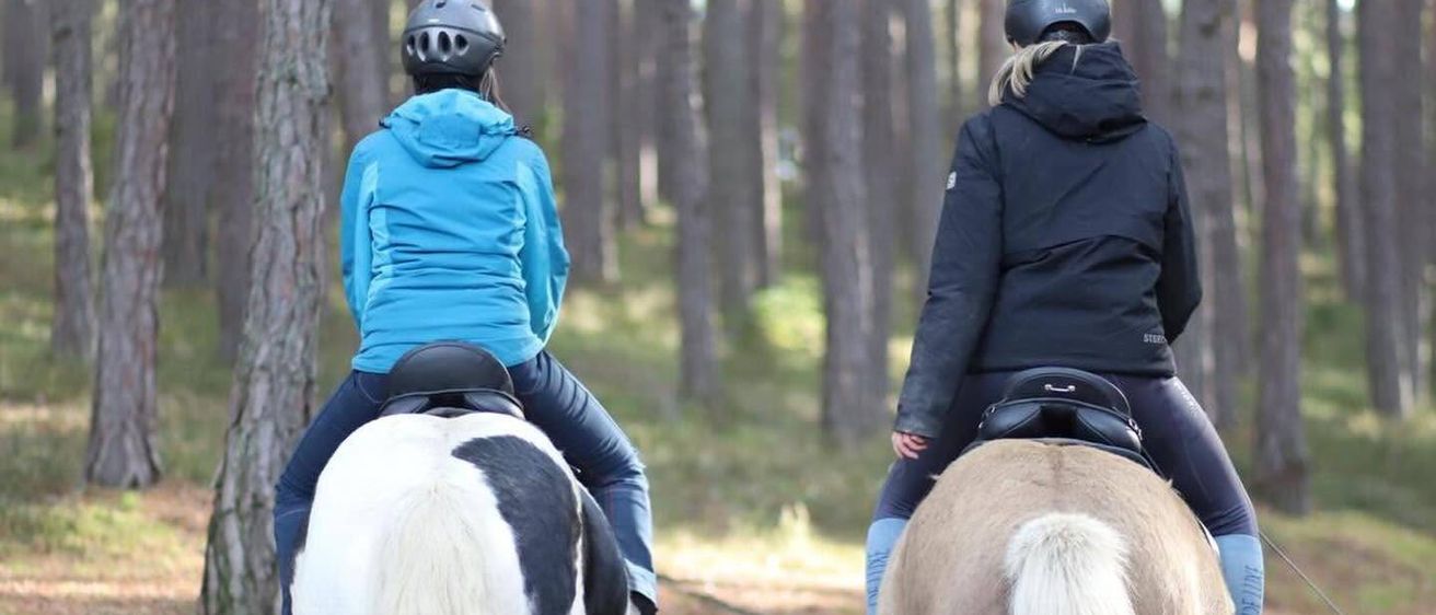 Zwei Personen reiten Pferde in einem Wald, beide tragen Helme und Jacken. Das Pferd links ist weiß und schwarz, das rechts ist hellbraun.