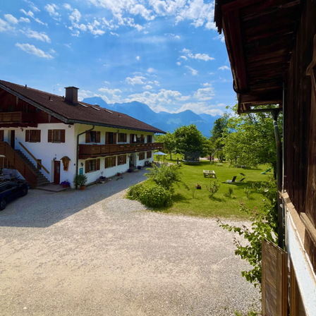 Ein traditionelles weißes und braunes Haus mit Balkonen, eingebettet in einem üppigen Garten mit Bergen im Hintergrund.