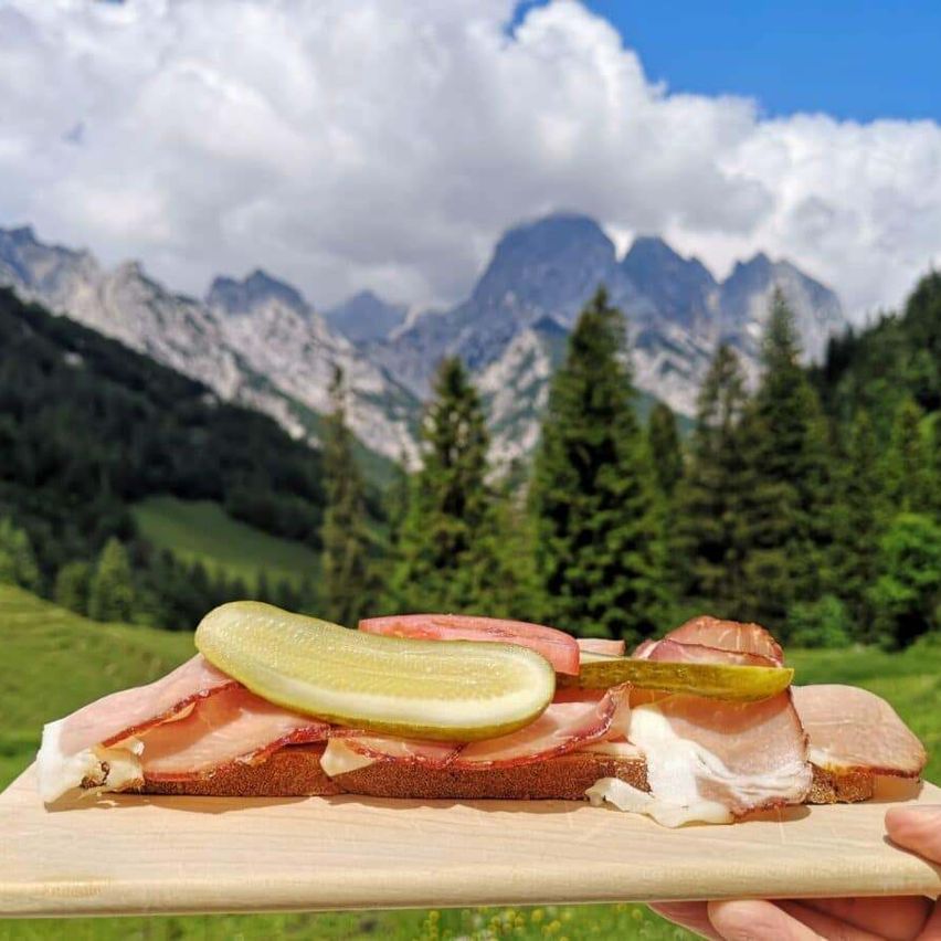 Ein Sandwich mit Schinken, Käse und Gurken auf einem Holzbrett, gehalten in einer Hand mit Bergen im Hintergrund.