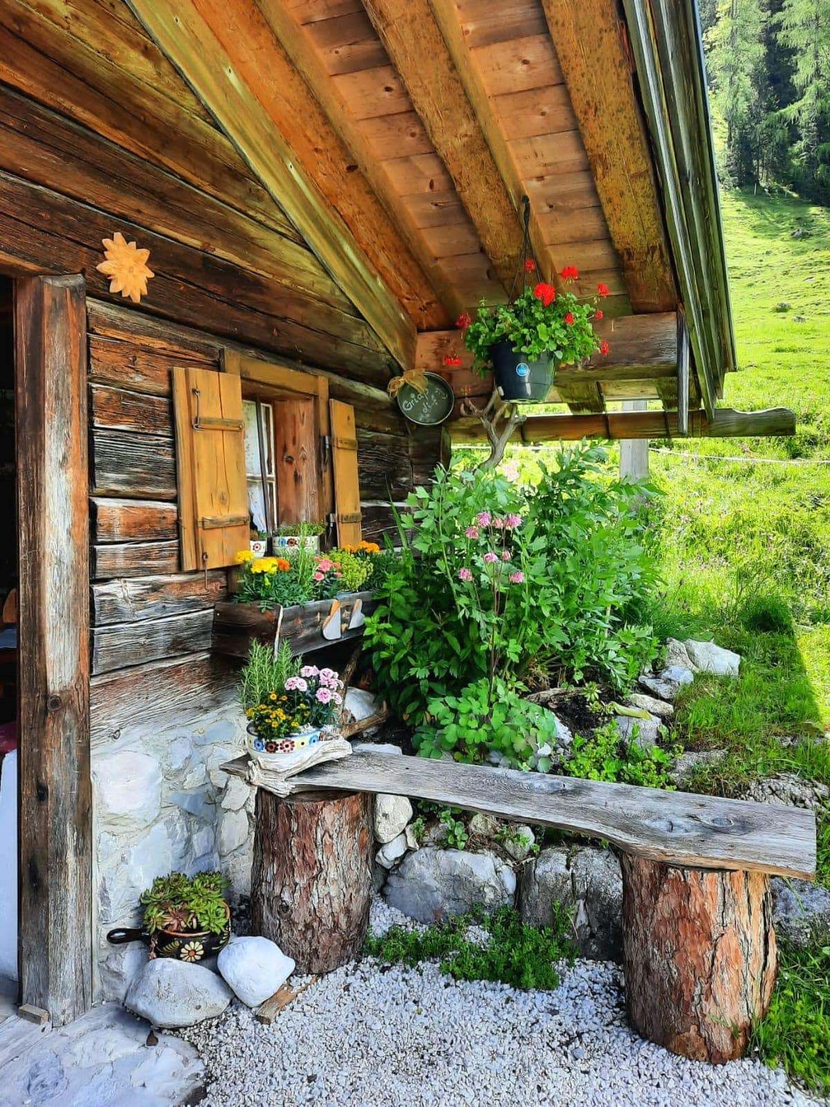 Eine Holzhütte mit blumengeschmücktem Fensterbrett und hängenden Blumentöpfen. Eine Bank und Topfpflanzen im vorderen Garten.