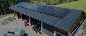 Eine Draufsicht auf ein Haus mit einer Solarpanelanlage auf dem Dach. Das Haus ist von grünen Bäumen und einer Bergkette umgeben. Ein getarntes Auto ist an der Seite geparkt.