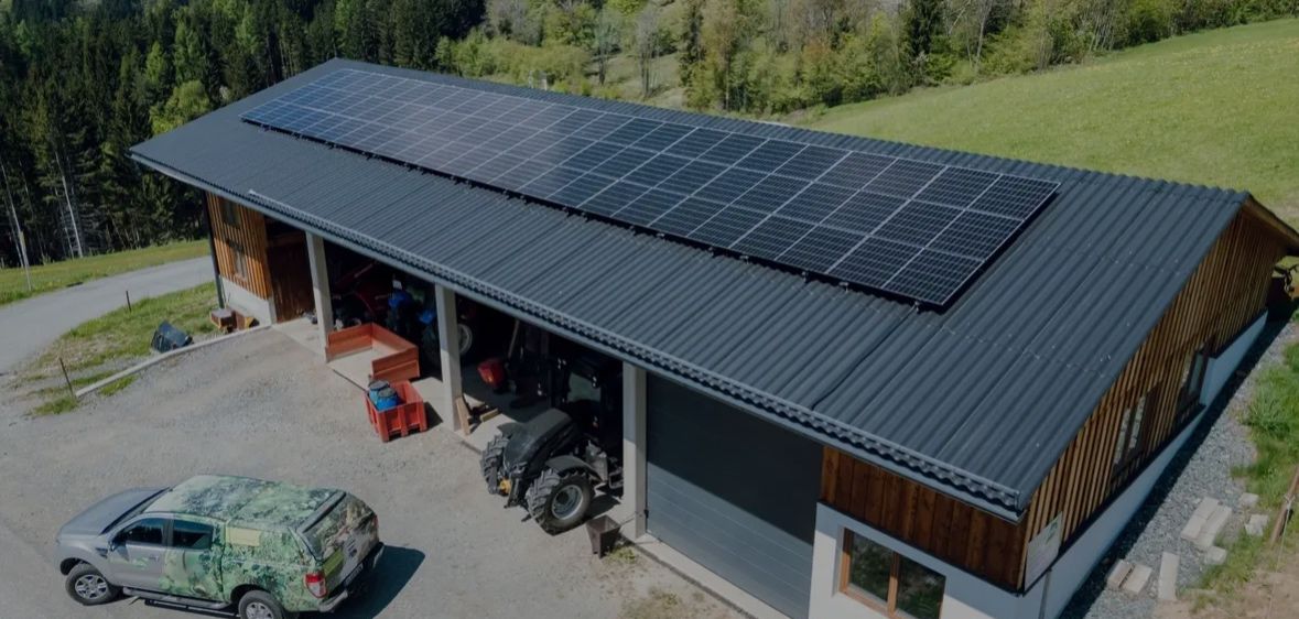 Eine Draufsicht auf ein Haus mit einer Solarpanelanlage auf dem Dach. Das Haus ist von grünen Bäumen und einer Bergkette umgeben. Ein getarntes Auto ist an der Seite geparkt.