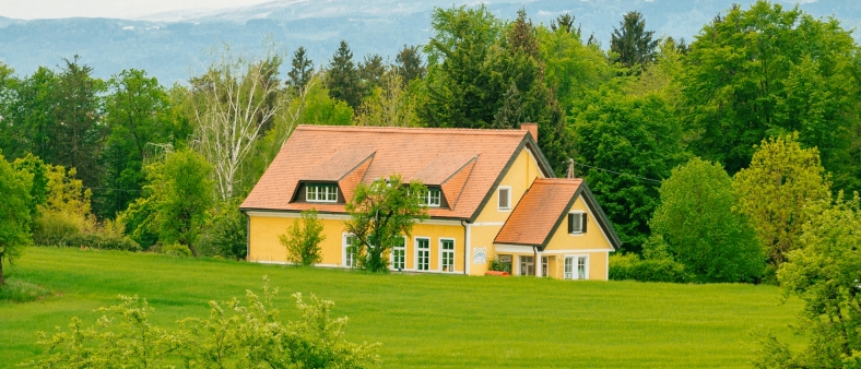 Ein gelbes Haus mit einem braunen Dach ist von üppigen grünen Bäumen und Büschen umgeben. Es befindet sich in einem weitläufigen grünen Feld mit entfernten Bergen.