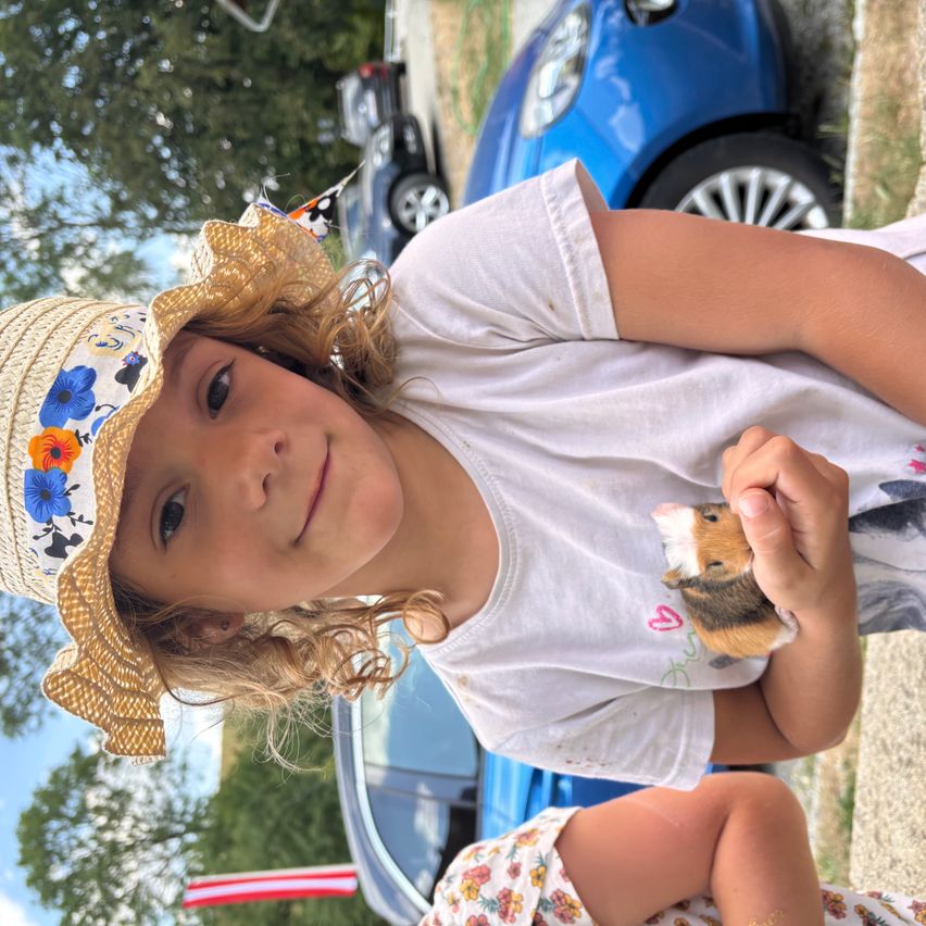A young girl wearing a straw hat with flowers holds a guinea pig in her hands. She is standing in front of a blue car.