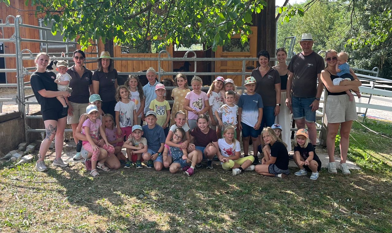 A group of children and adults are posing for a photo in a grassy area with a tree and a wooden fence in the background. Some of the children are wearing hats.