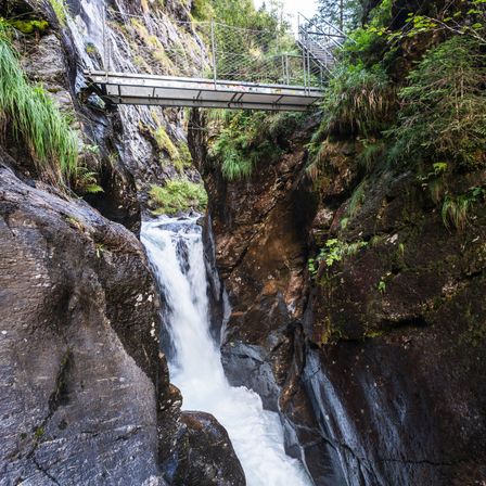 Ein Wasserfall stürzt zwischen zwei felsigen Klippen hinab, mit einer kleinen Brücke darüber. Der Wasserfall ist von üppigem grünen Bewuchs umgeben.