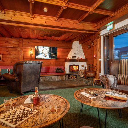 Ein gemütlicher Raum mit Holzwänden und einem Kamin. Zwei Sessel stehen sich gegenüber um einen Schachbrett auf einem runden Tisch. Ein Fernseher ist an der Wand montiert und ein Glasfenster bietet einen Blick nach draußen.