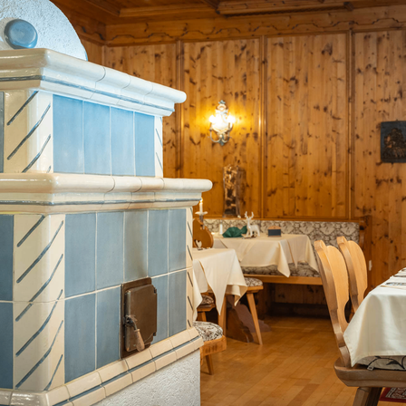 Ein gemütliches Restaurant mit Holzwänden, das einen gefliesten Ofen, eine Lampe an der Wand und einen Tisch mit einem weißen Tuch aufweist. Stühle sind um den Tisch herum angeordnet. Eine Holzbank steht an der Wand. Ein gerahmtes Bild ist an der Wand angebracht.