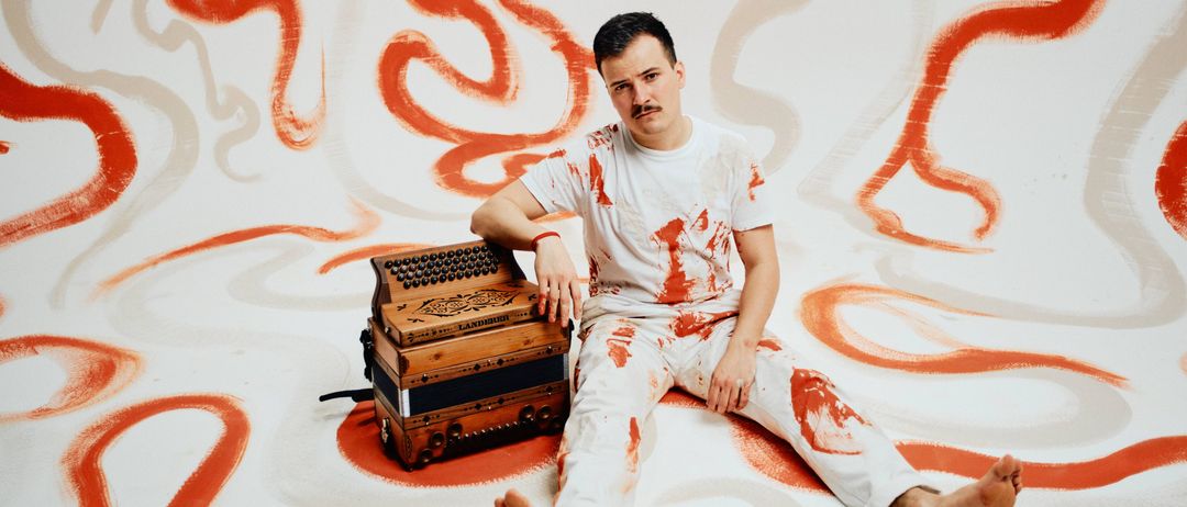 Ein Mann in einem weißen T-Shirt und Hose sitzt auf einer weißen Oberfläche mit roten Farbspritzern und hält ein Akkordeon.