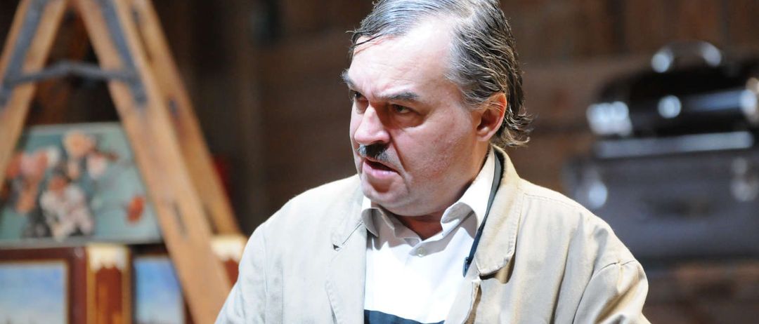 Ein Mann mit grauem Haar und einem Schnurrbart sieht ernst aus, während er vor hölzernen Malstaffeln steht.