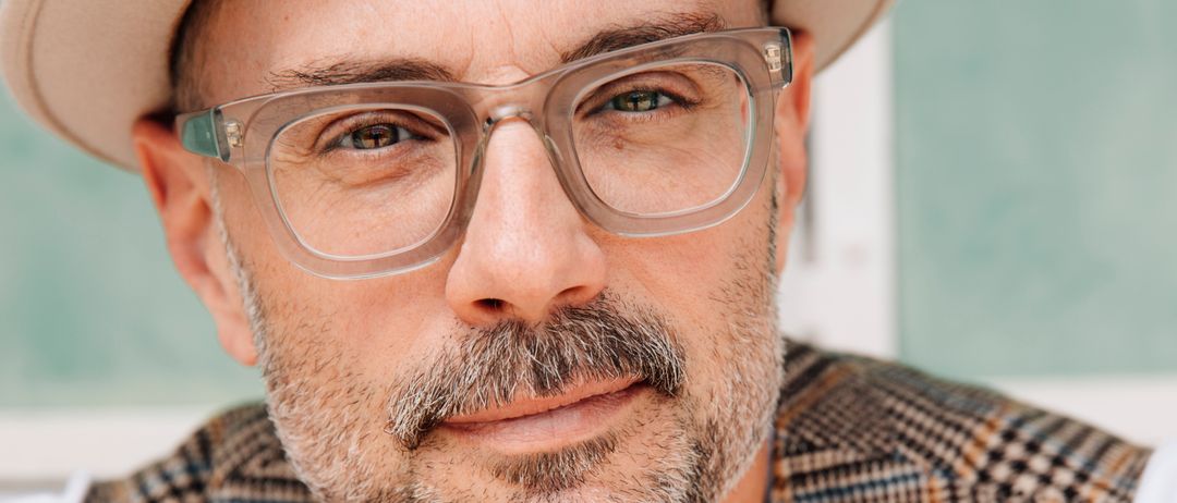 Ein Mann mit Bart und Schnurrbart trägt eine Mütze und eine Brille und lächelt in die Kamera. Er ist in einer karierten Weste über einem weißen T-Shirt gekleidet.